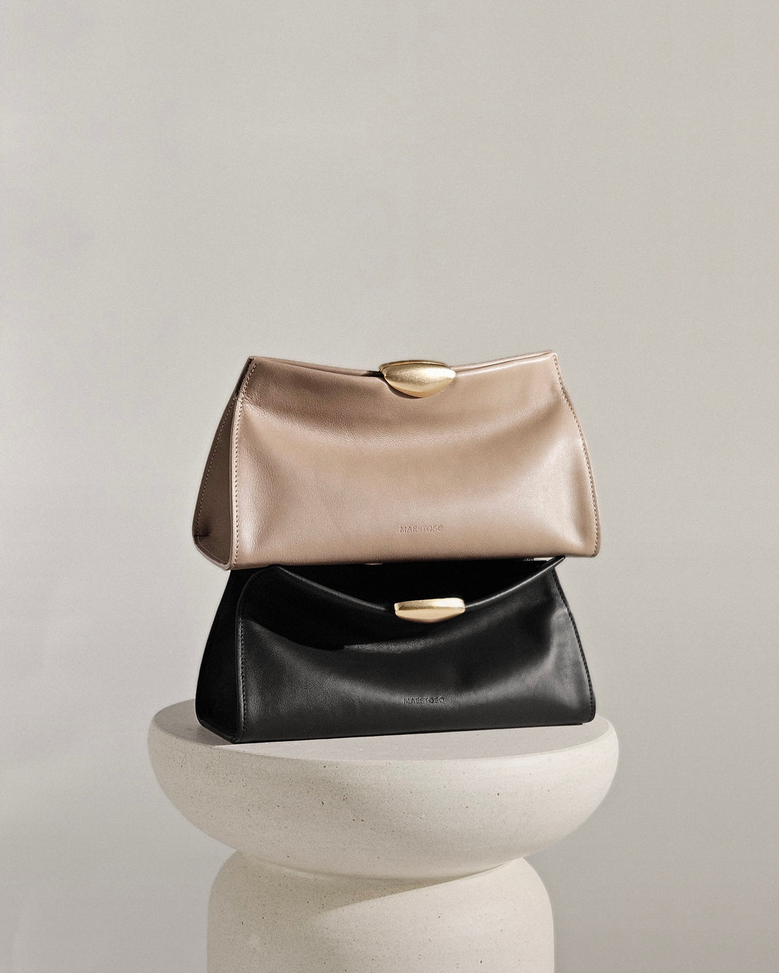 Two Maison Maestoso Noa Soft handbags, one black and one beige, on a white pedestal against a light gray background.