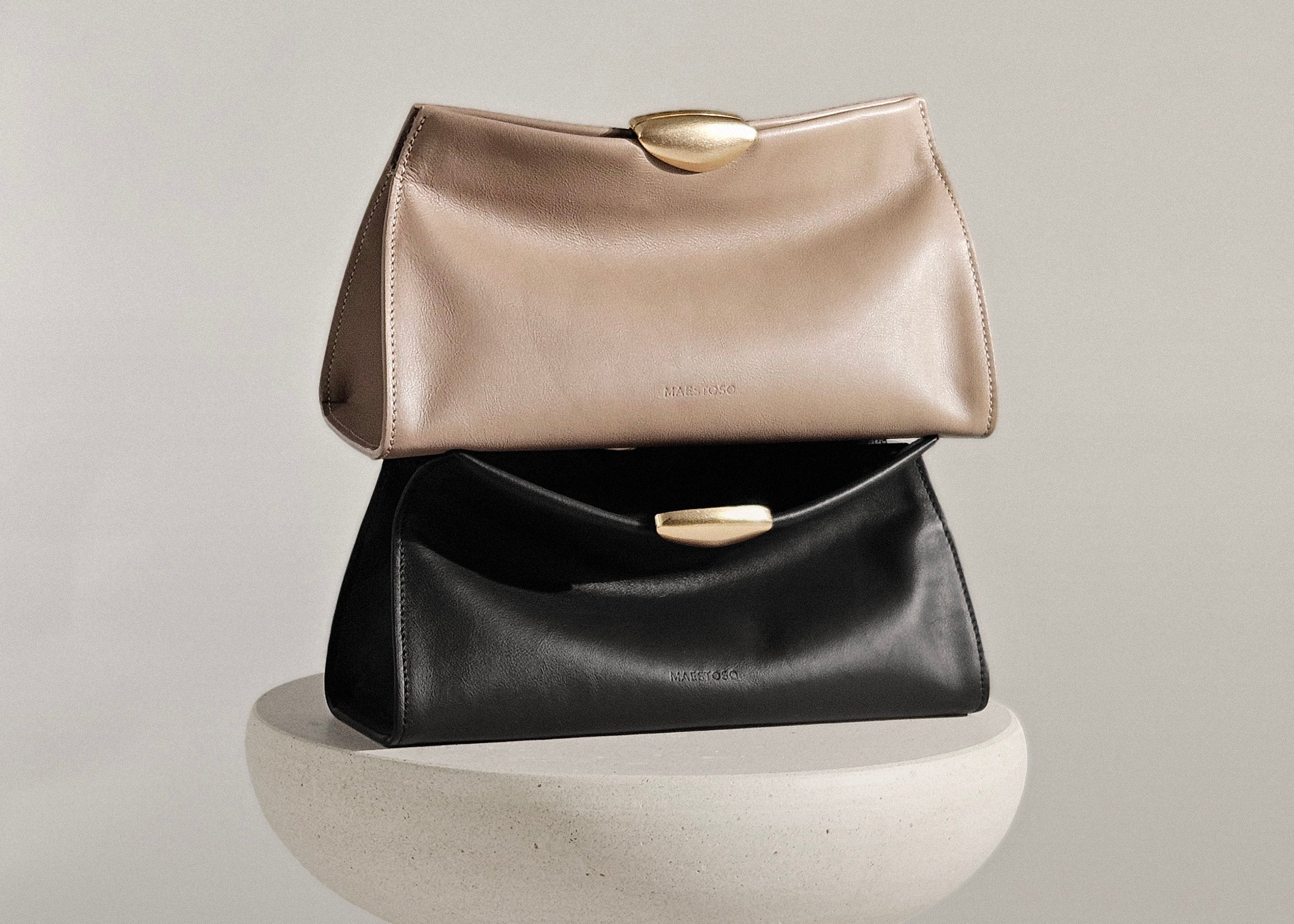 Two Maison Maestoso Noa Soft leather handbags stacked on a round pedestal against a light gray background. The top bag is a warm champagne beige with a gold clasp, and the bottom bag is black with a matching gold clasp.